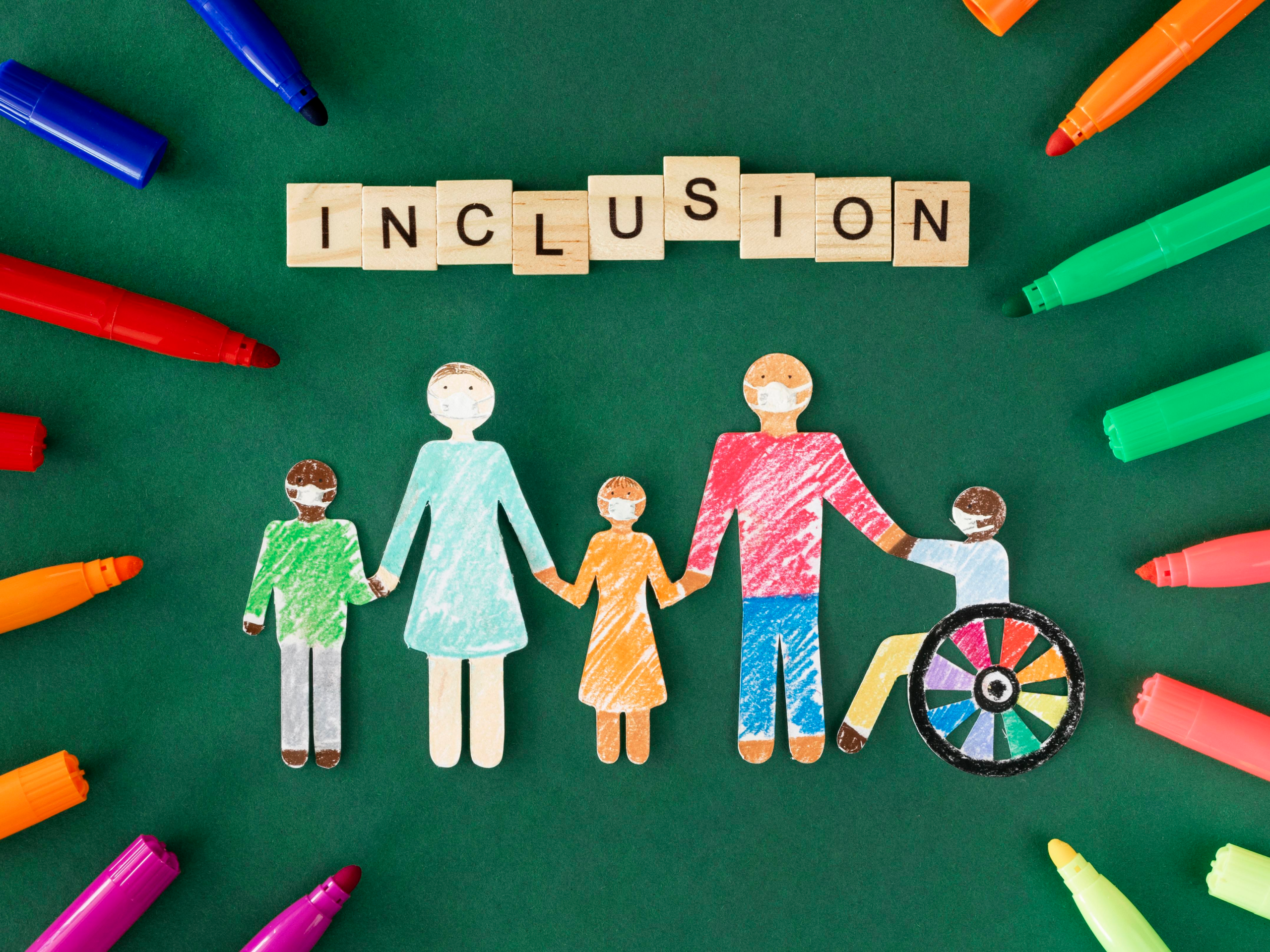 Ilustración de figuras de papel representando a una familia diversa con la palabra “INCLUSION” escrita en bloques de madera y rodeada de marcadores de colores.