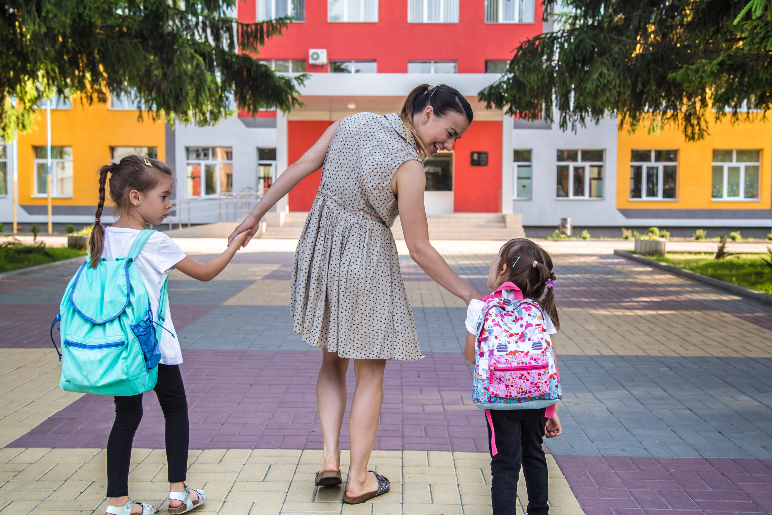 Madre acompañando a dos niñas con mochilas coloridas hacia la entrada de una escuela de fachada moderna y colorida.