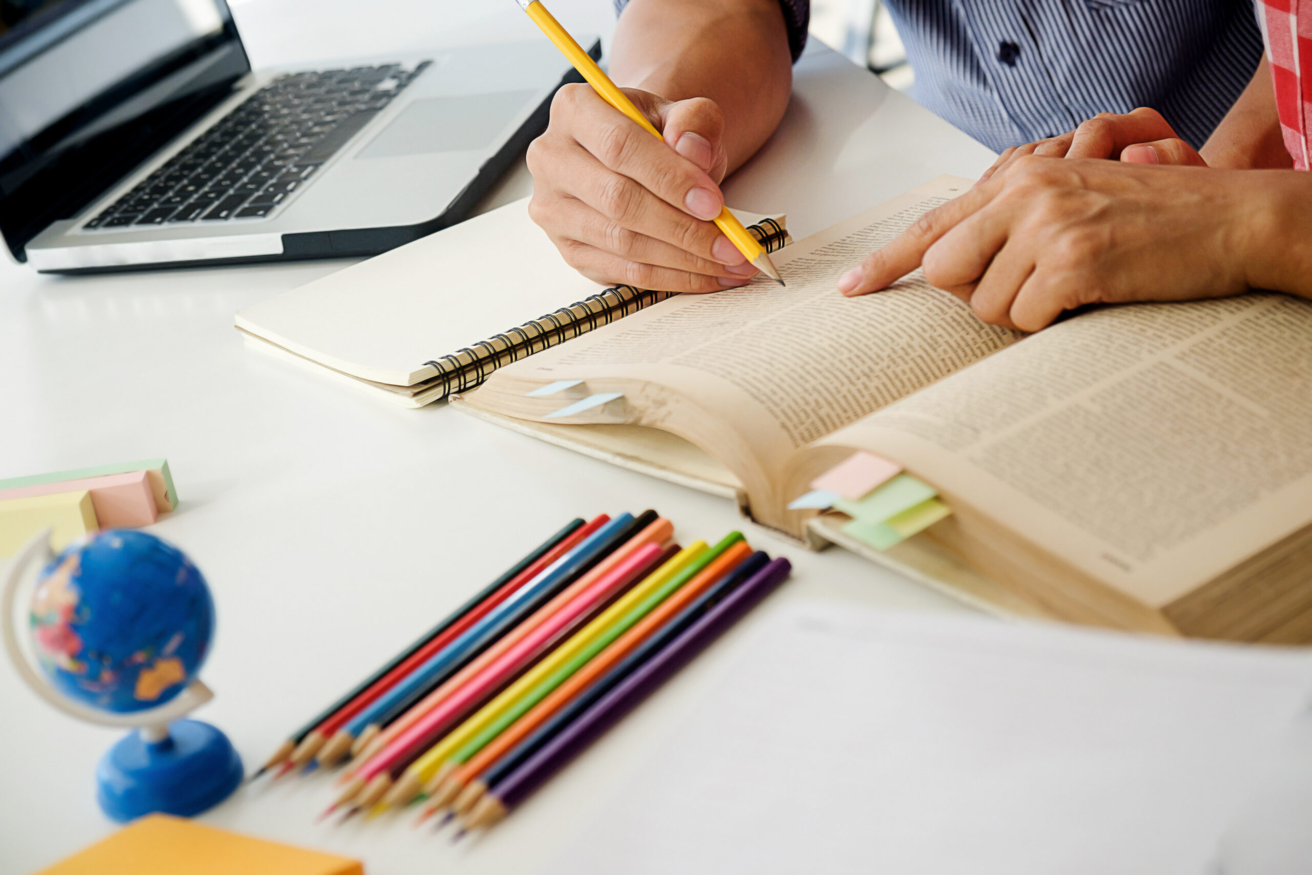 Personas estudiando juntas con un libro abierto, cuaderno, lápiz, computadora portátil y lápices de colores sobre una mesa blanca.