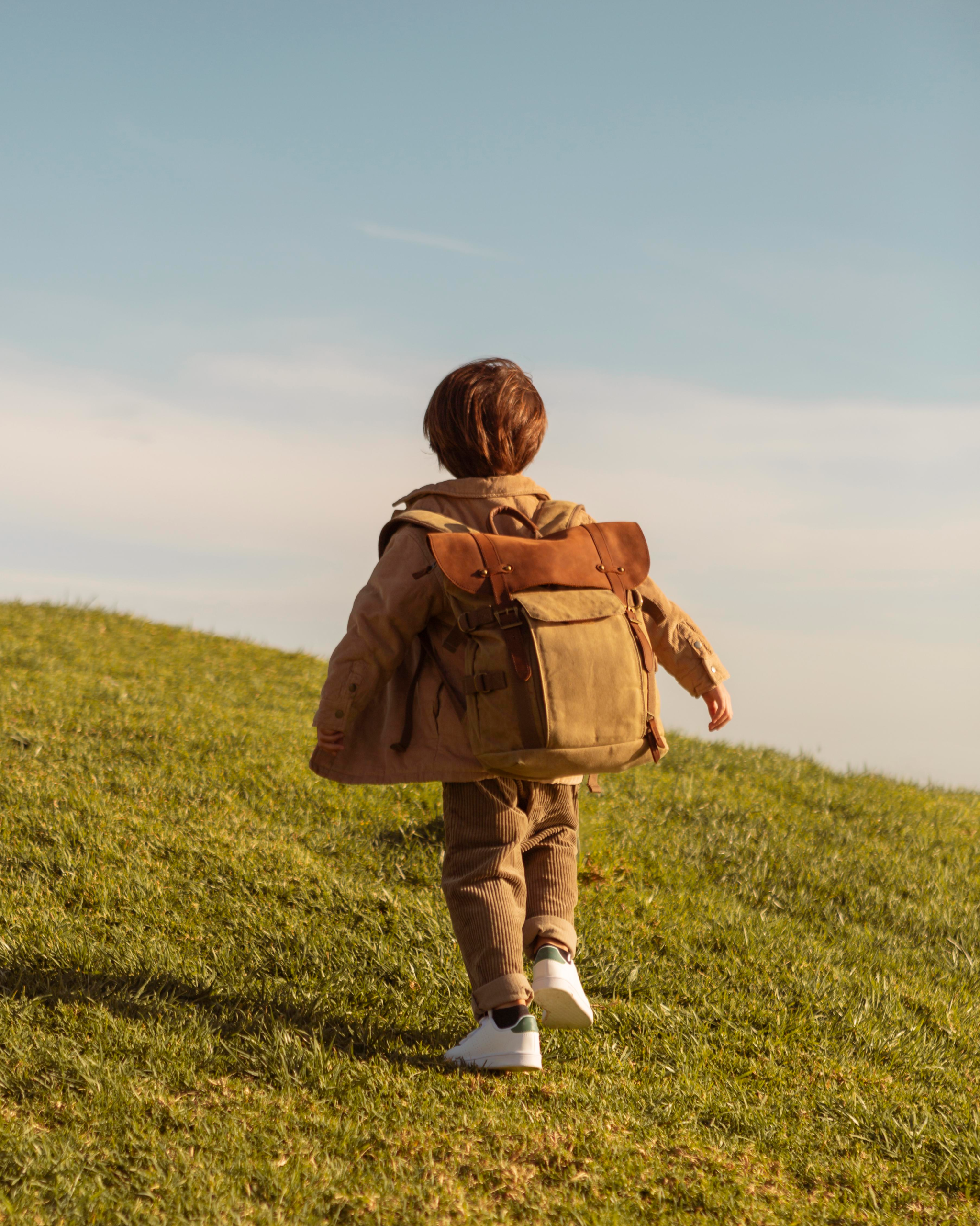 Niño con mochila caminando sobre una colina verde bajo un cielo despejado.