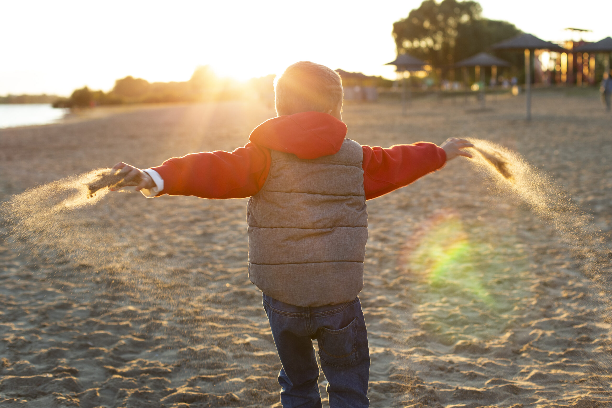 Niño jugando con arena en la playa durante el atardecer, con los brazos extendidos.