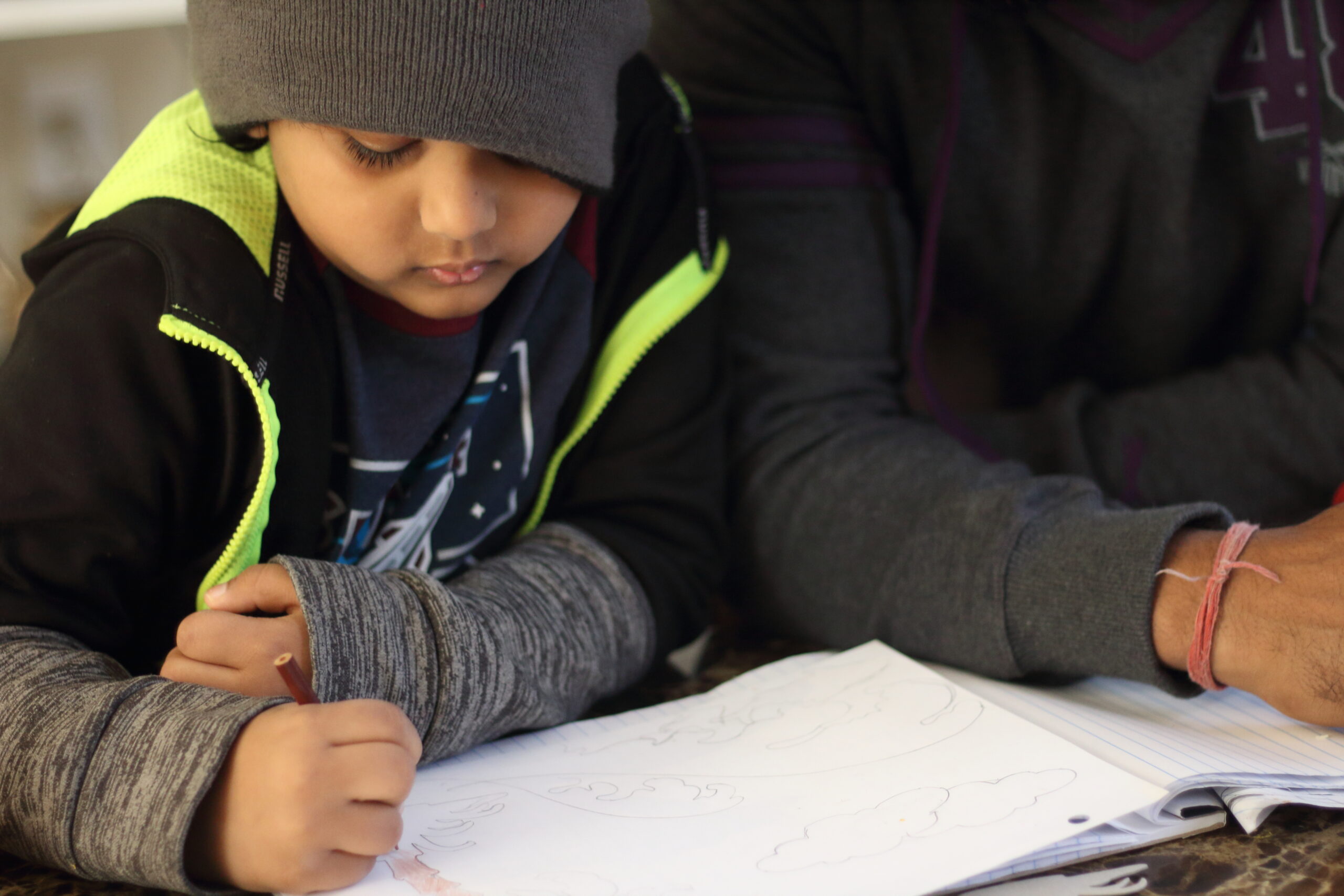 Un niño con gorro dibuja en un cuaderno mientras su padre lo acompaña y supervisa.