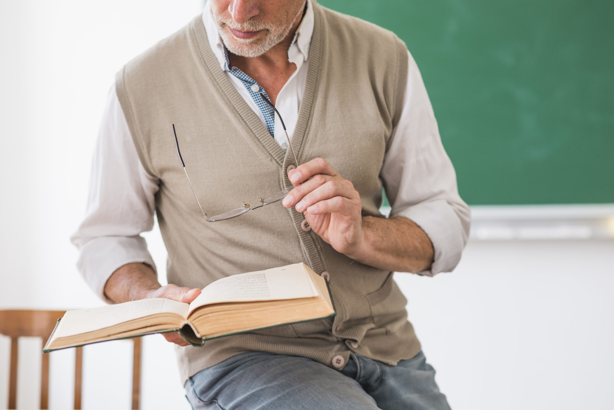 Profesor adulto sosteniendo un libro abierto y unos anteojos frente a un pizarrón en el aula.
