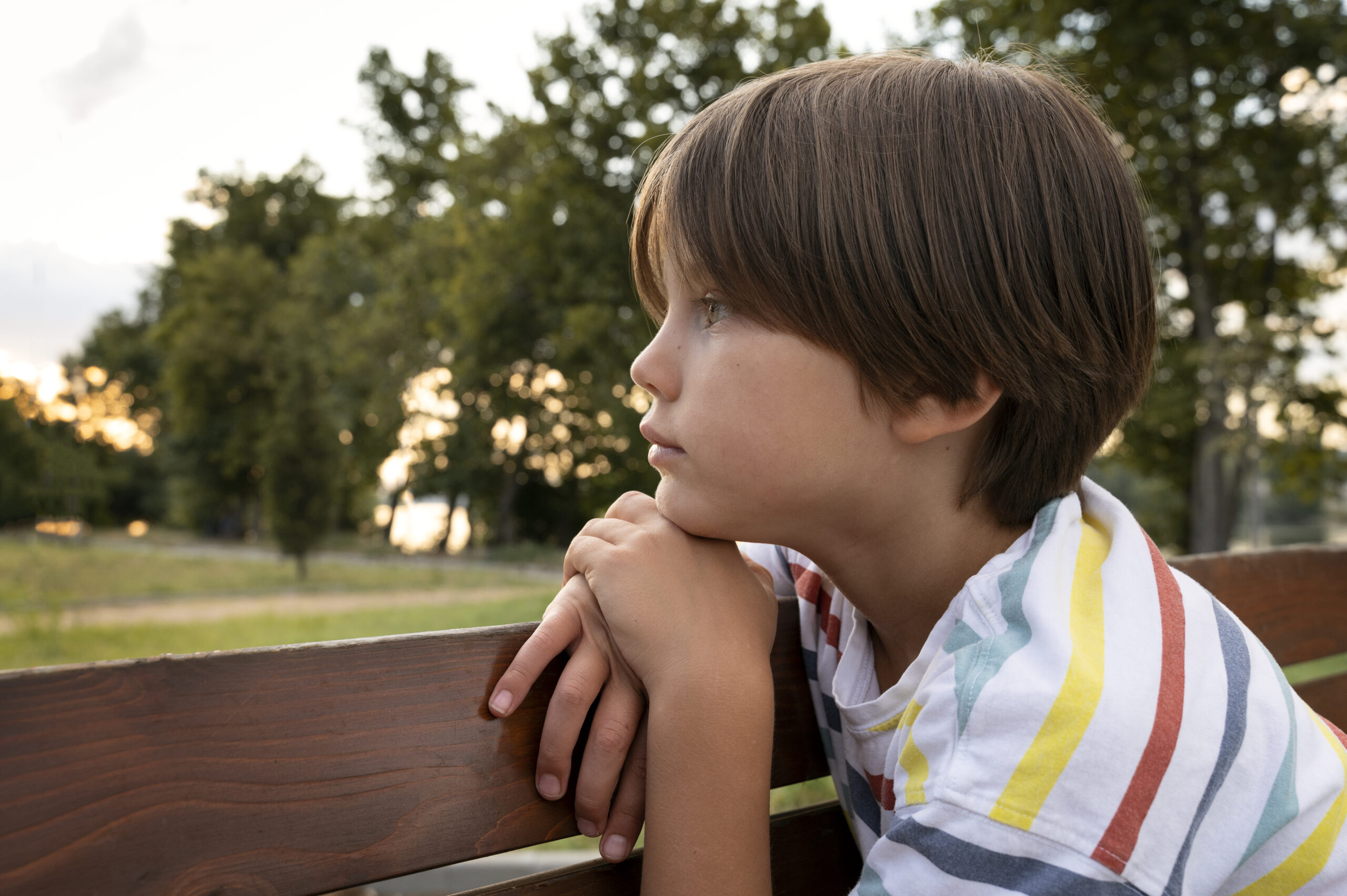 Niño sentado en un banco de madera observando el horizonte durante el atardecer, con expresión pensativa.