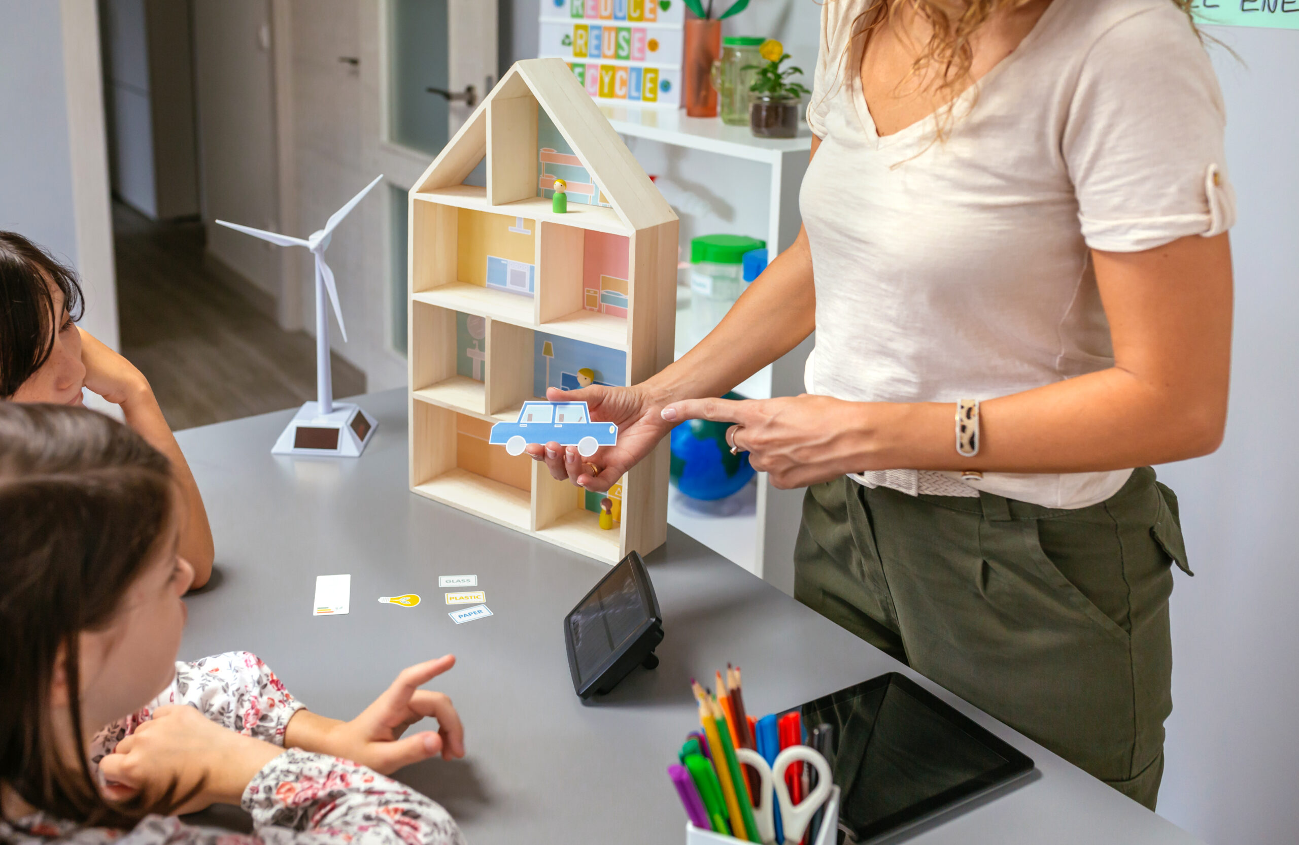 Docente mostrando a sus alumnos una maqueta de casa sustentable y un modelo de auto eléctrico en una clase sobre energía renovable.